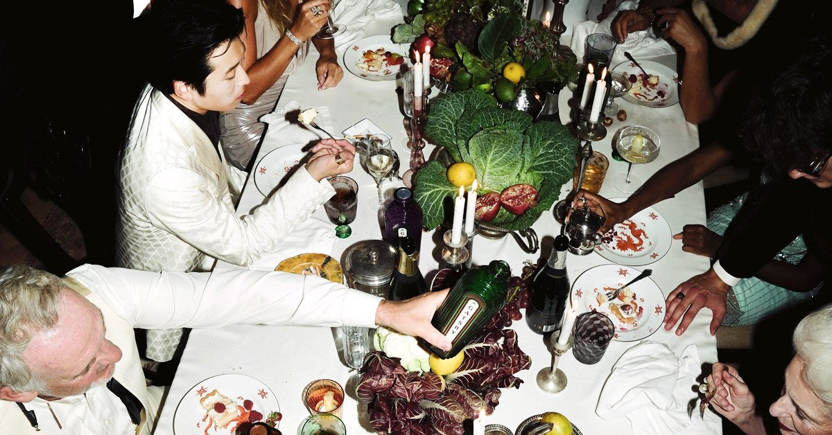 A candlelit dinner party seen from above, a bottle of Gatsby Gin passed across a long table of glasses, candles and fruit.