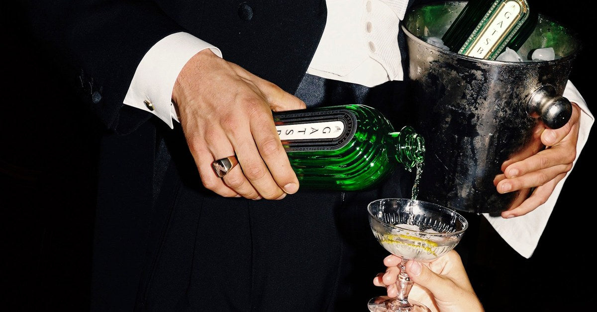 A man in white tie pours Gatsby Gin into a coupe held out by a guest, a second bottle chilling in a silver ice bucket.
