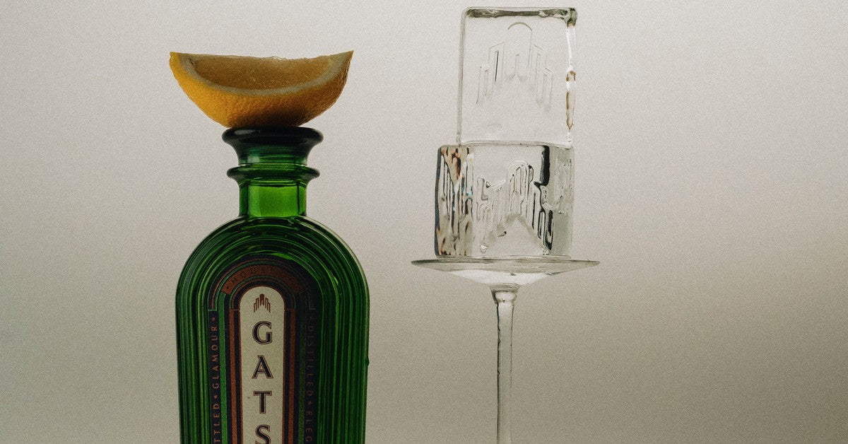 A bottle of Gatsby Gin topped with a lemon wedge beside a martini glass holding a block of ice.