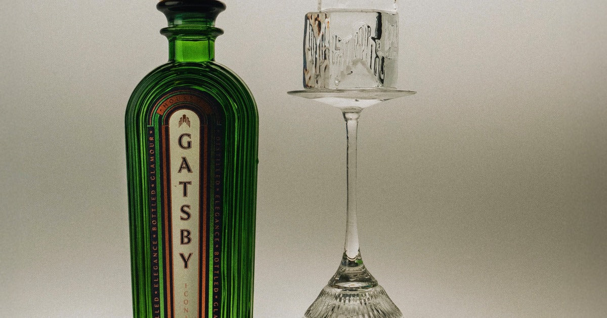 A bottle of Gatsby Gin beside an upturned coupe glass holding ice, on a silver tray in warm studio light.