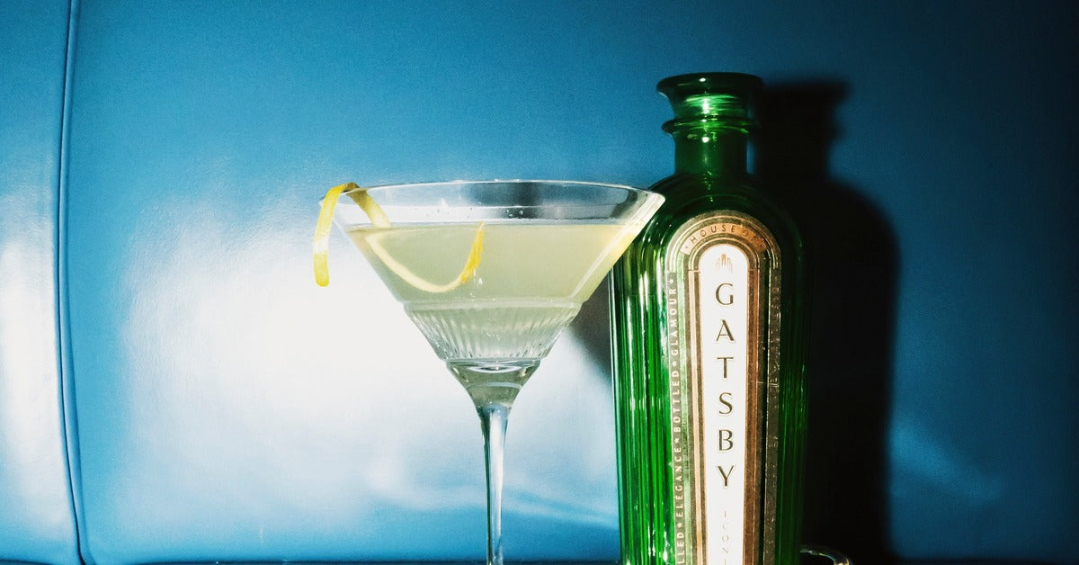 A gin martini with a lemon twist beside a bottle of Gatsby Gin on a silver tray, against blue leather.