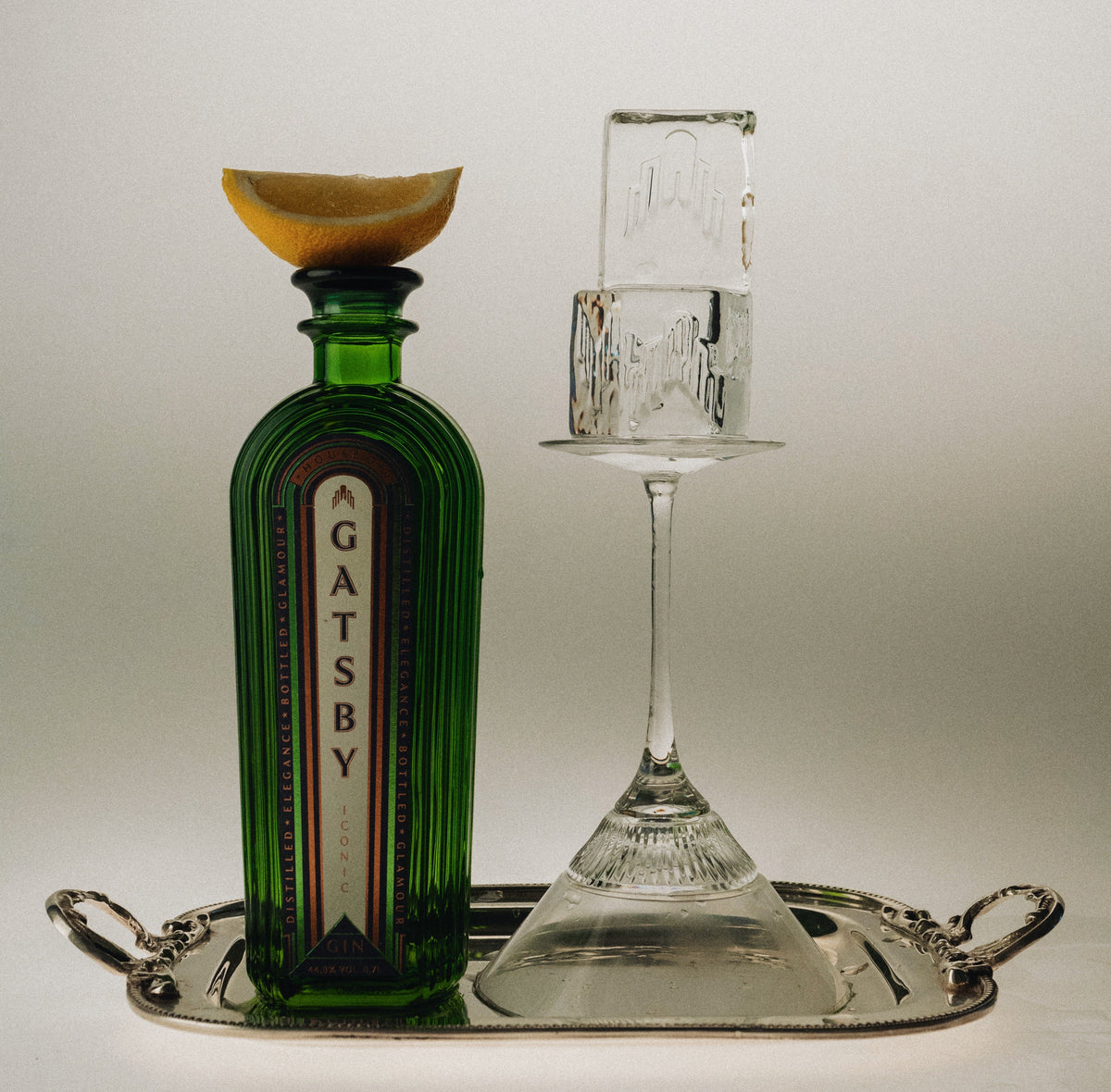 A bottle of Gatsby Gin on a silver tray beside a coupe glass and a block of ice